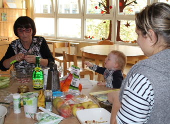 Auf dem Bild ist ein Kind mit zwei Erwachsenen an einem Küchentisch beim Salatzubereiten zu sehen