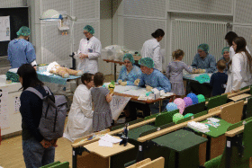 In einem Raum im Krankenhaus mit vielen Erwachsenen und einigen Kindern wird auch ein Teddy operiert.