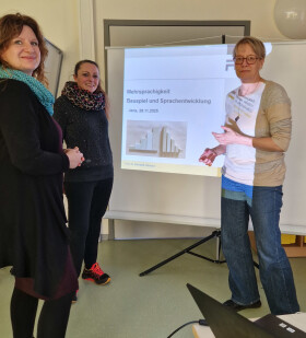 3 Frauen stehen vor einer Beamer Leinwand und sind im Gespräch über die dahinter zusehende Überschrift das stattgefundenen Vortrags