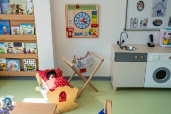 Puppenecke im Kindergarten mit Puppenwiege mit einer zugedeckten Puppe, an der Wand eine Uhr mit einem Kalender, daneben eine kleiner Wäscheständer mit Puppenwäsche 