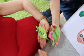 Mutter mit selbstgebastelter Raupe auf einem ausgeschnittenen Papier-Blatt auf der Hand, daneben die Hand des Kindes mit einer weiteren selbstgebastelten Raupe sua Pfeifenputzern. Aush hier sitzt die Raupe auf einem grünen Blatt, welches aus Papier ausgeschnitten wurde