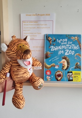 Dokument an weißer Wand berichtet über Zahnprojekt, daneben ein Zahputzbuch und ein Plüsch-Zahn-Putz-Tiger